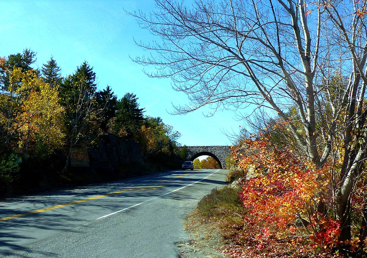 A stone arch bridge crossing the Park Loop Road in autumn, with a car passing beneath it and red and yellow foliage on the roadside
