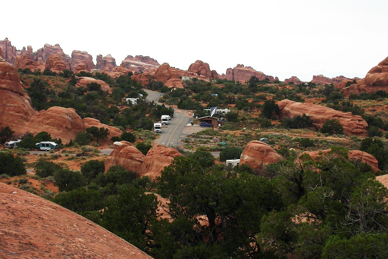 An elevated view over Devils Garden Campground showing small RVs and tents scattered among red rock outcrops and juniper