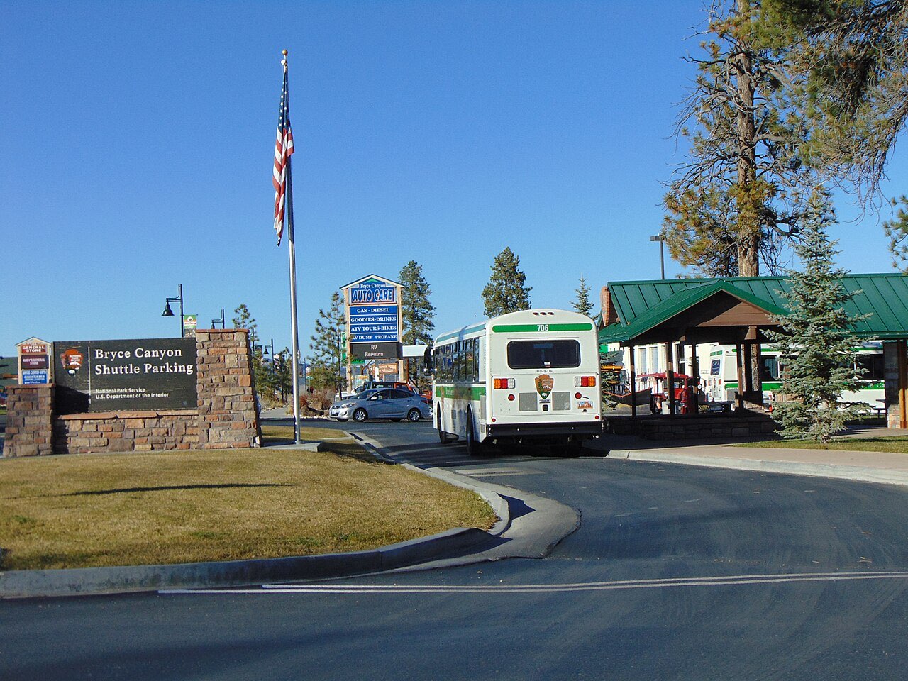 A green and white Bryce Canyon shuttle bus at the Shuttle Parking stop in Bryce Canyon City, with a stone sign and flagpole