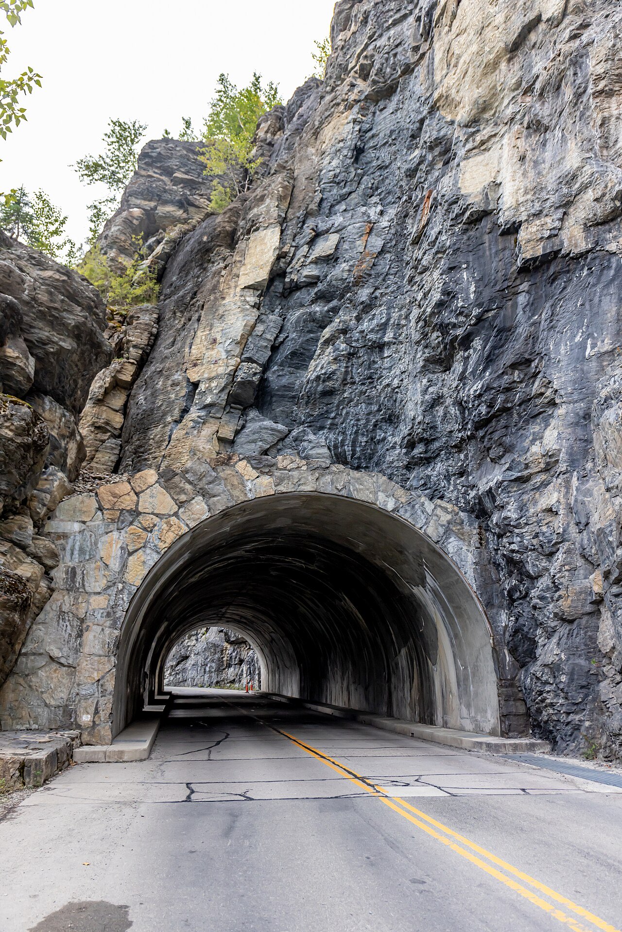 Narrow stone-arched tunnel portal on Going-to-the-Sun Road cut through a dark rock face, two-lane road with a double yellow line