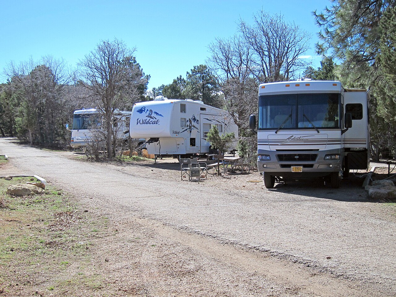 A Class A motorhome and a fifth-wheel trailer parked on adjacent gravel sites at Trailer Village, with trees between the sites