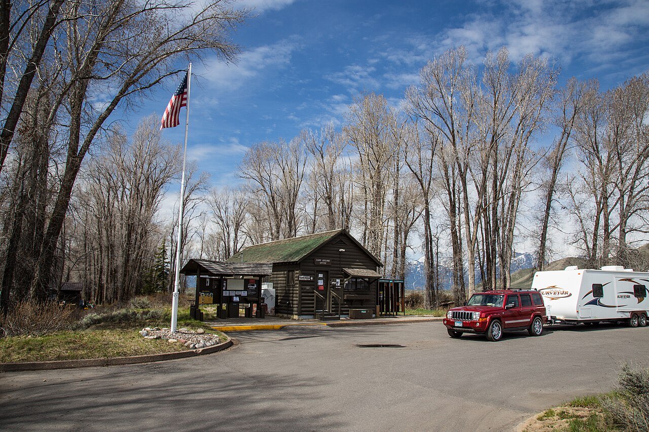 Log campground office at Gros Ventre Campground with a red SUV towing a white travel trailer parked beside it under bare cottonwoods