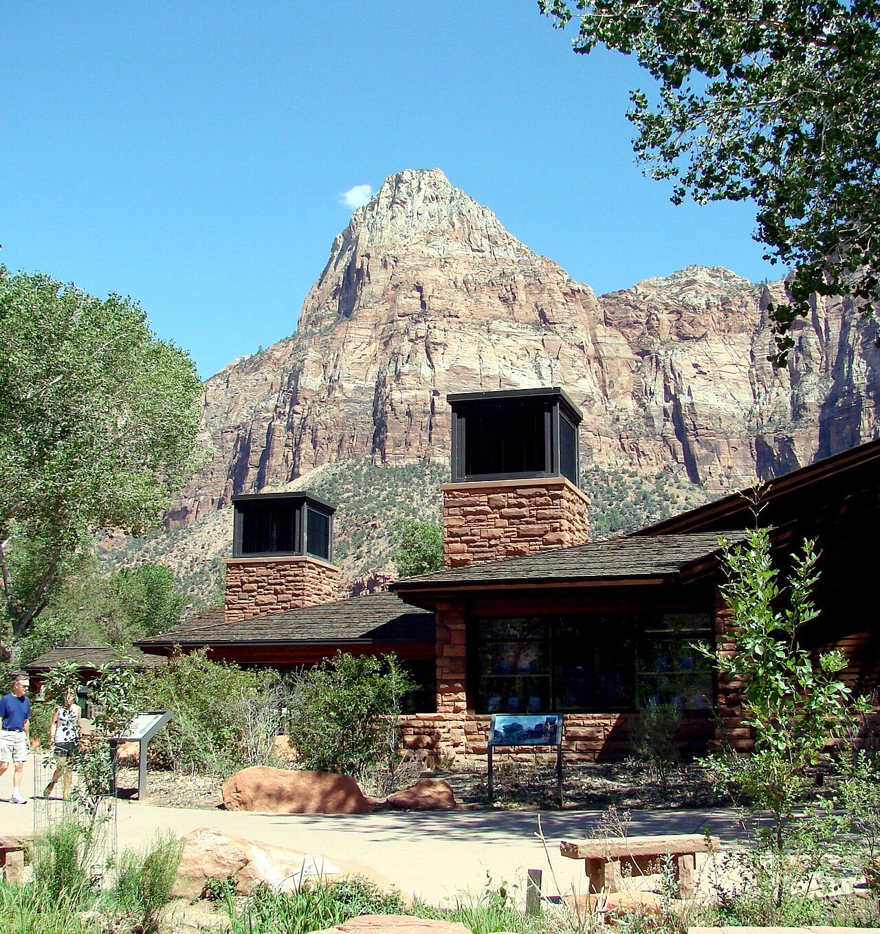 Stone and timber Zion Canyon Visitor Center building with a sandstone peak rising behind it under a blue sky